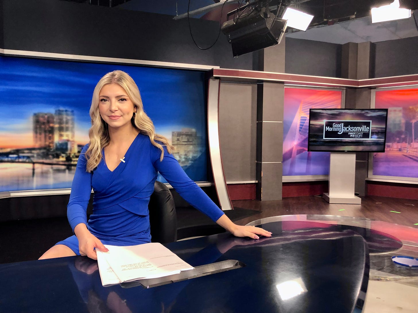A blonde woman in a blue V-neck dress sits at a television anchor desk. A faint scar is visible on her chest.