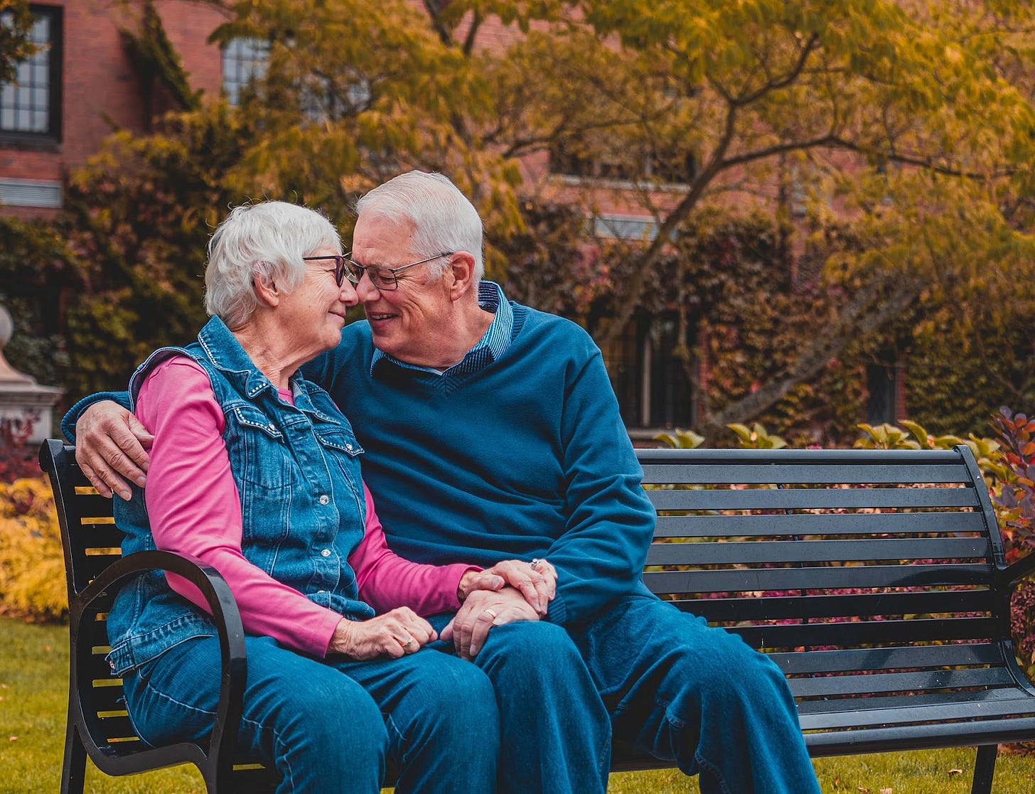 A grey haired couple touching noses on a bench.