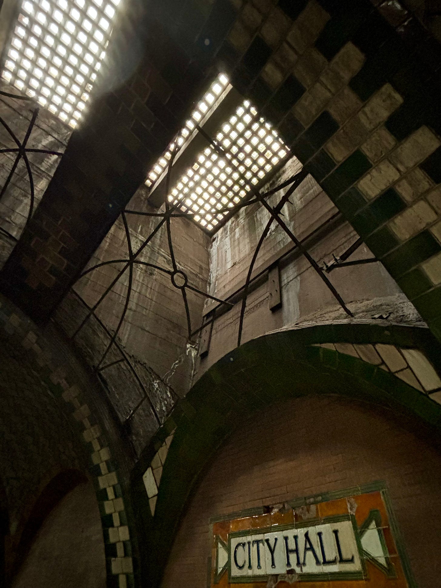 The wall of a disused subway station labeled City Hall. Above it is an arched ceiling with a skylight. There has been decorative metal sculpted into an intricate design but most of the metal is gone now.