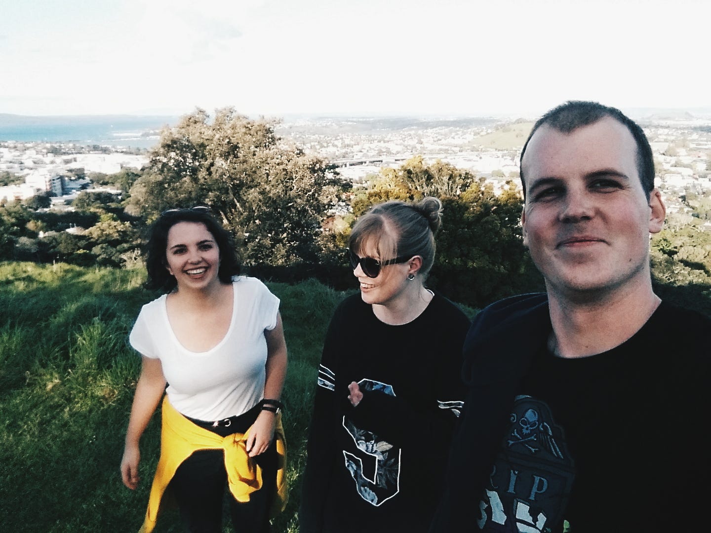 A selfie that Lachie is taking, mid-way up Mt Eden, looking out across Auckland. Louise is on the left, Chelsea in the middle, and Lachie on the right. Louise and Lachie are looking at the camera, while Chelsea is looking left. They're all smiling and laughing. 