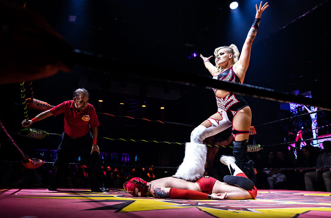 A female wrestler stnads above her vanquished opponent