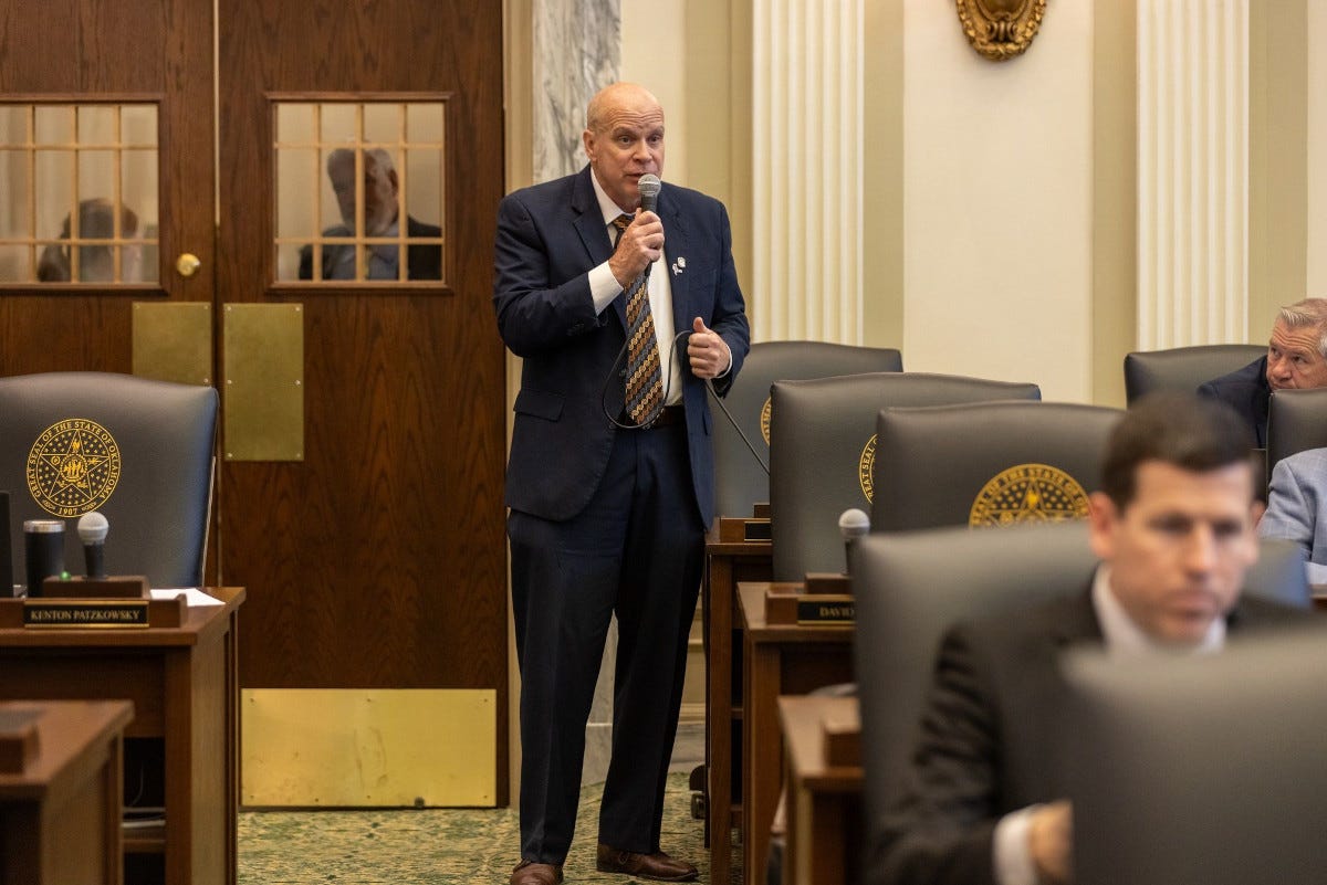 Man holding a microphone and wearing a blue suit addresses a group a lawmakers. Man holding a microphone and wearing a blue suit addresses a group a lawmakers.