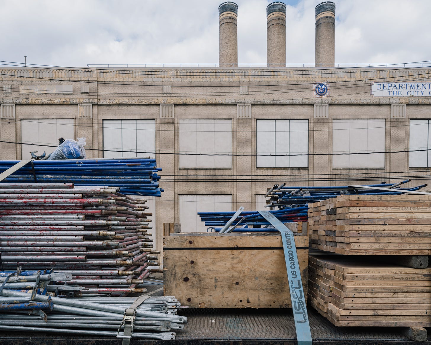 Stacks-Of-Metal-Pipes-And-Wooden-Planks-In-Foreground-With-Three-Yellow-Brick-Smokestacks-Behind