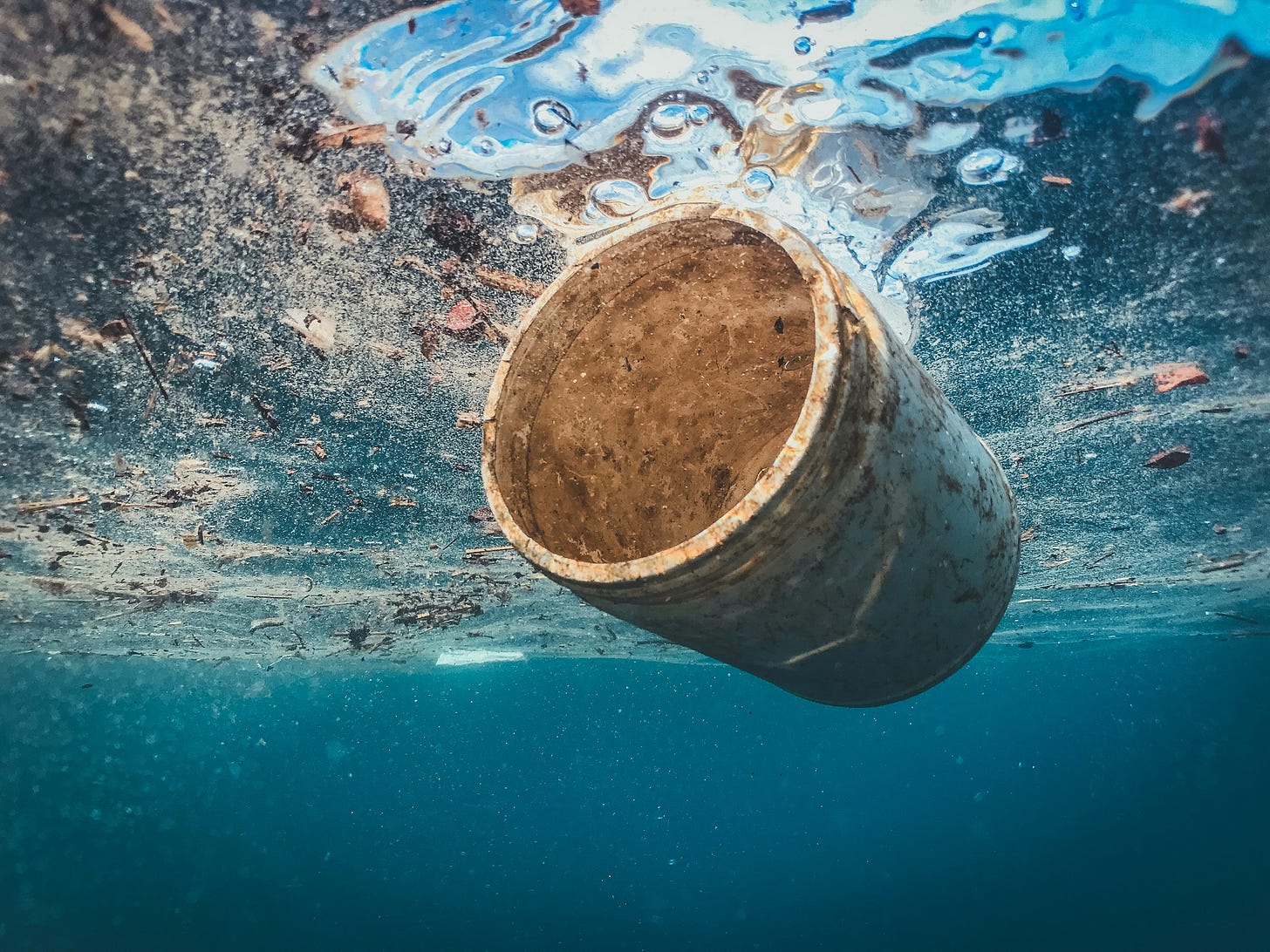 Vista marina pochi metri sotto la superficie del mare, con in primo piano un tubo di plastica incrostato e tutt'attorno altri pezzettini di plastica e sporco che galleggiano.