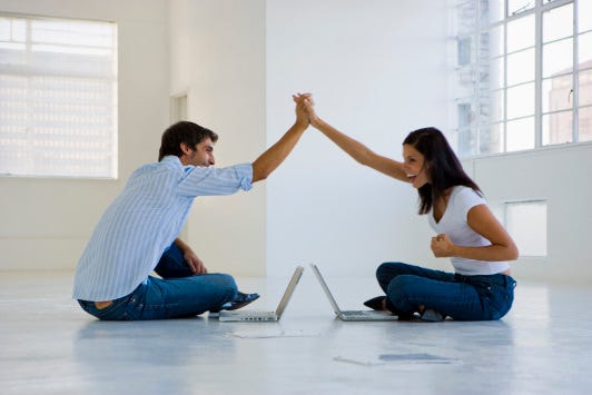 Man on left and woman on right sitting cross legged on floor facing each other over open laptops while giving each other a high five.