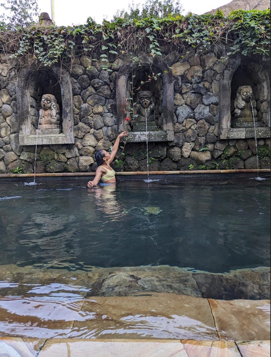 Nathalie is in a pool in Bali. There are 3 statues by where she stands. She extends her arm to reach for a red flower that's hanging from a branch