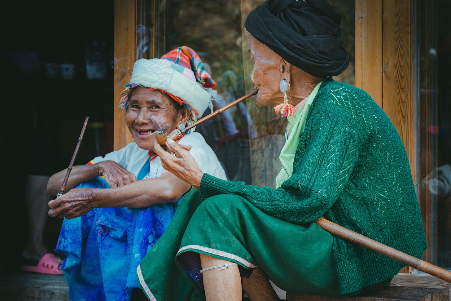 Duas idosas em trajes típicos do leste ou sudeste asiático, sentadas à porta de uma casa, têm longos cachimbos nas mãos. Uma delas olha para a câmera e sorri.