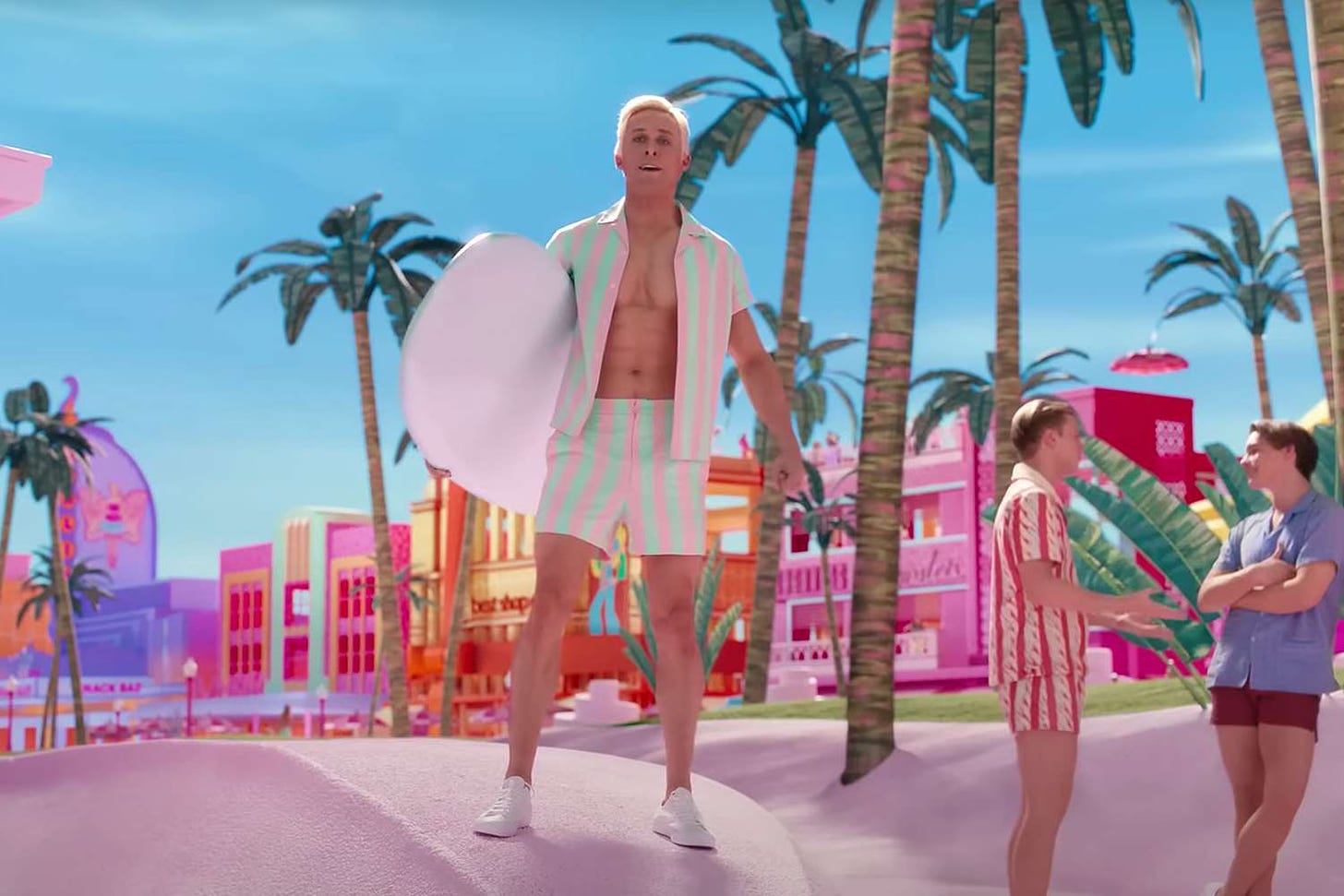 Ryan Gosling as Ken in Barbie, standing at the edge of the beach carrying a surfboard.