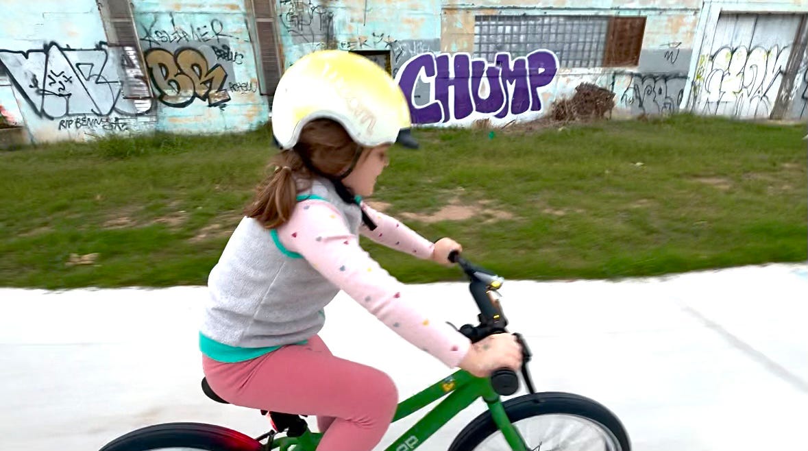 Child riding new bike past industrial wall covered in graffiti, including big purple "CHUMP"