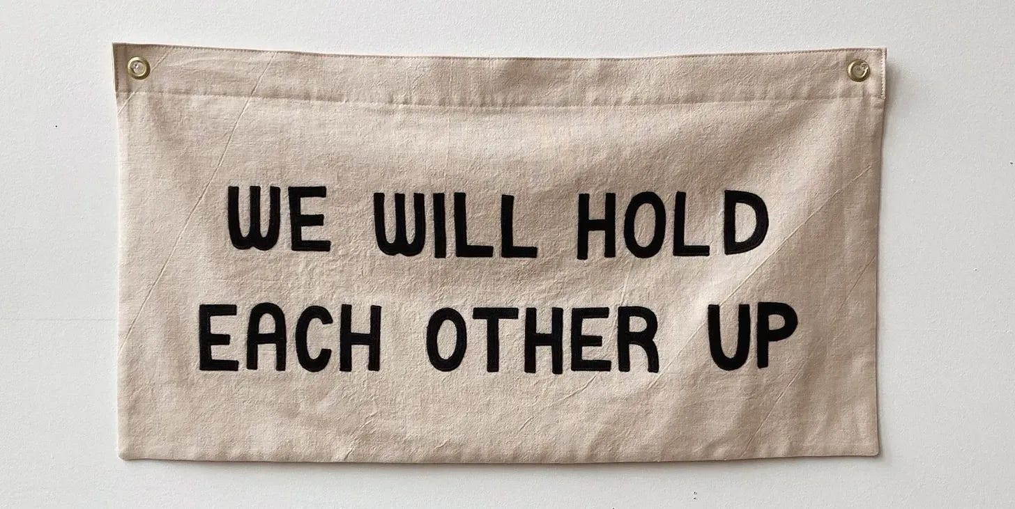Fabric wall hanging with bold black text reading “WE WILL HOLD EACH OTHER UP.” The banner is beige with metal grommets in the top corners. Artist credit: Ashley Brown Durand. Fabric wall hanging with bold black text reading “WE WILL HOLD EACH OTHER UP.” The banner is beige with metal grommets in the top corners. Artist credit: Ashley Brown Durand.