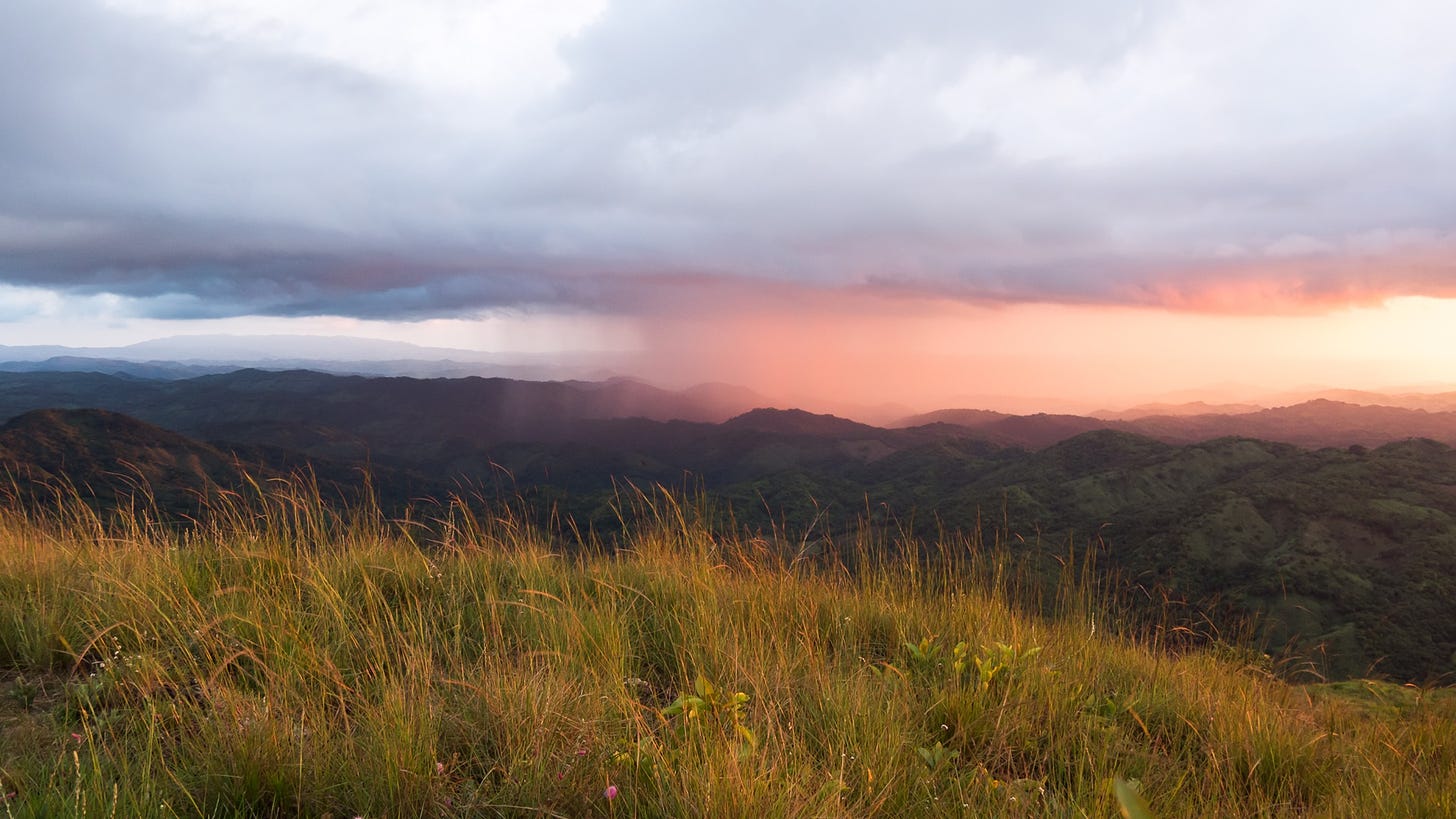 A grassy hill in the foreground and mountains in the background over which rain is falling.