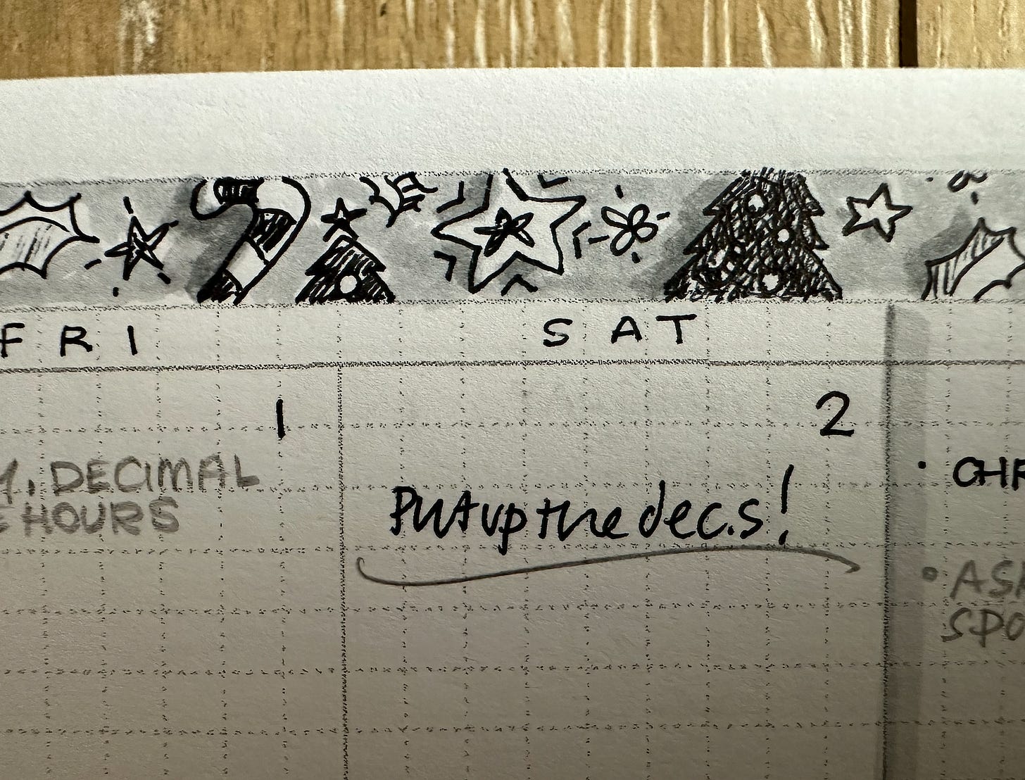 Closeup of a monthly calendar page, showing part of Friday and Saturday. There are rough black and grey drawings of various Christmas decorations in a banner across the top, including holly, a candy cane, stars, and a Christmas tree.