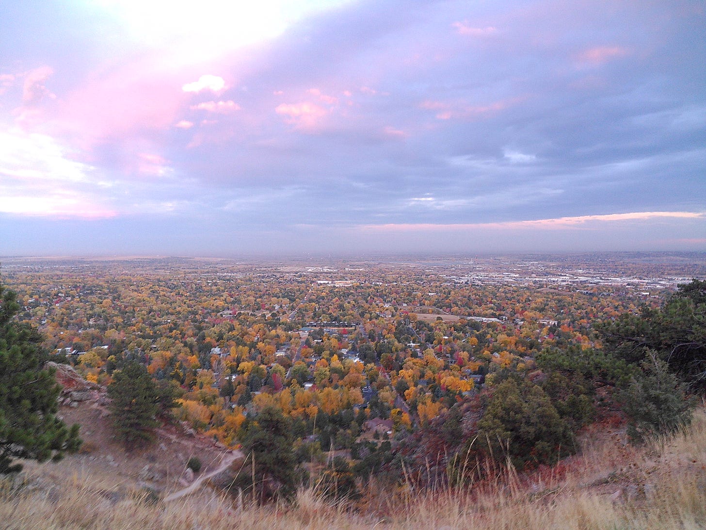 Fall foliage in Boulder, Colorado, as seen from the hiking trails of Mt Sanitas. Yellow, orange, and green trees in the foreground; a swirl of blues and purples in the sky.