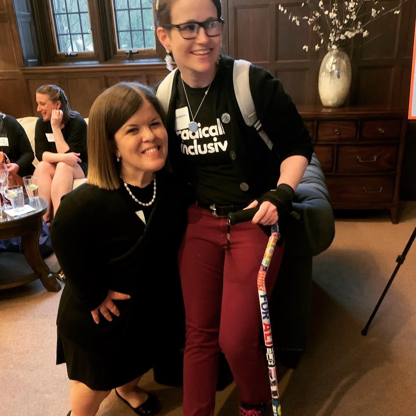 Two white women smiling. Woman 1 is a little person woman 2 is the author, brown hair, glasses,and cane. Her shirt says "radically inclusive"