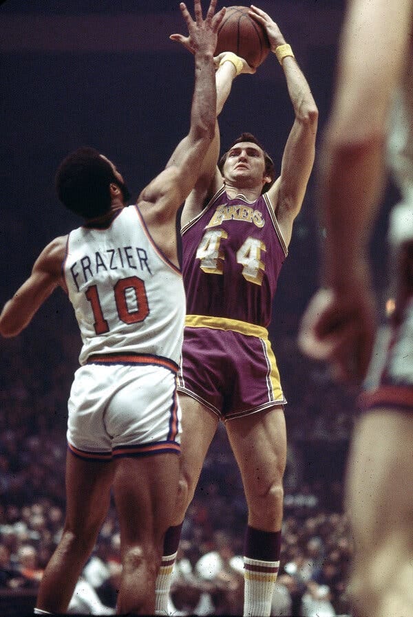 Jerry West, wearing a purple and hold Lakers uniform with the number 44, holds a basketball with both hands and prepares to shoot. Walt Frazier, wearing a white and orange Knicks uniform with the number 10, reaches up to block his shot.