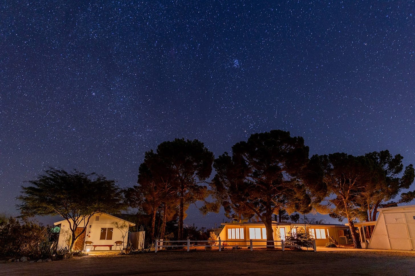 Starry nighttime shot of front exterior view of Jackrabbit Studios, including its Casita on the left, main house in the center, and edge of the studio workshop on the right, with a crown of midnight-blue sky dotted with billions of white stars.