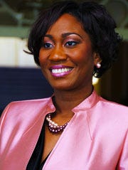 A woman in a pink suit jacket, with earrings and pearl necklaces, smiling at the camera 
