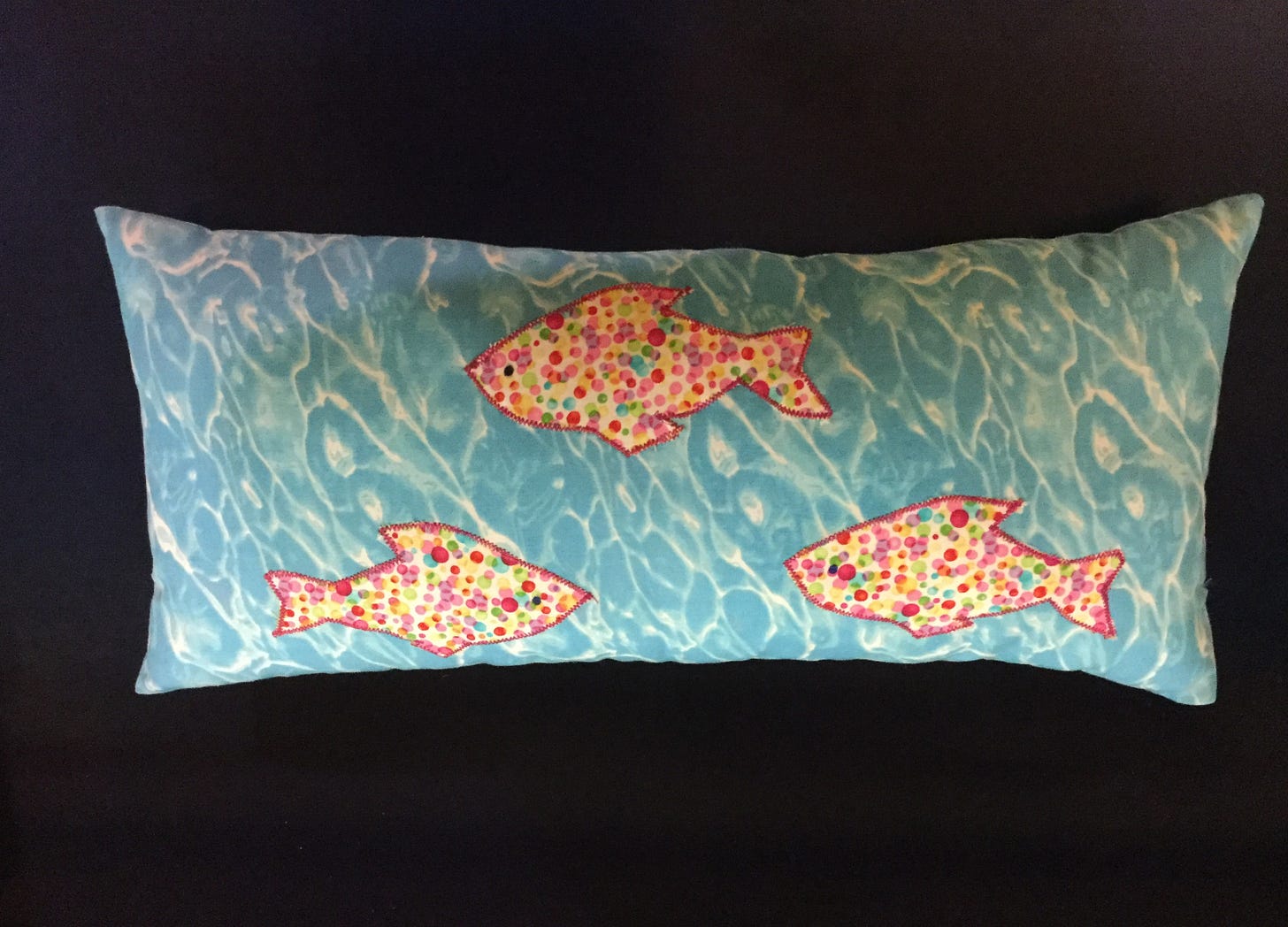 A small, narrow pillow made from fabric that looks like sunlit water and includes three appliquéd spotted fish.