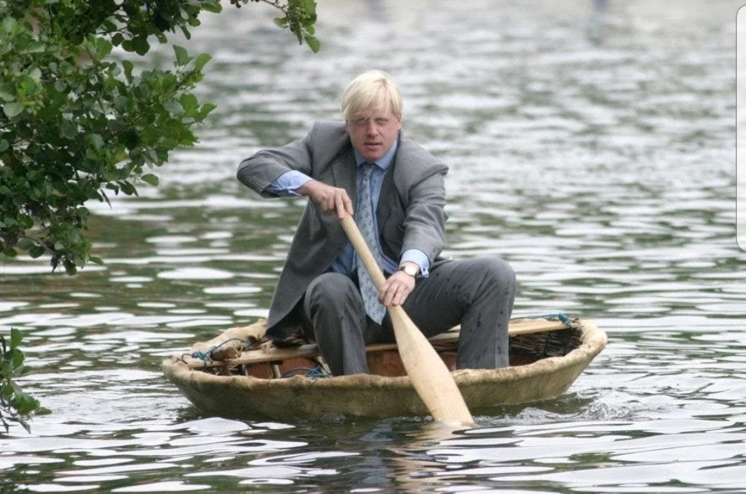 Image of a blonde middle aged man in a suit, inside a rowing boat, probably on a lake, rowing with just one paddle