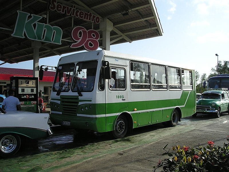 A boxy green and white bus is seen at a gas pump in Cuba while a line of buses and vintage cars can be seen in the background.