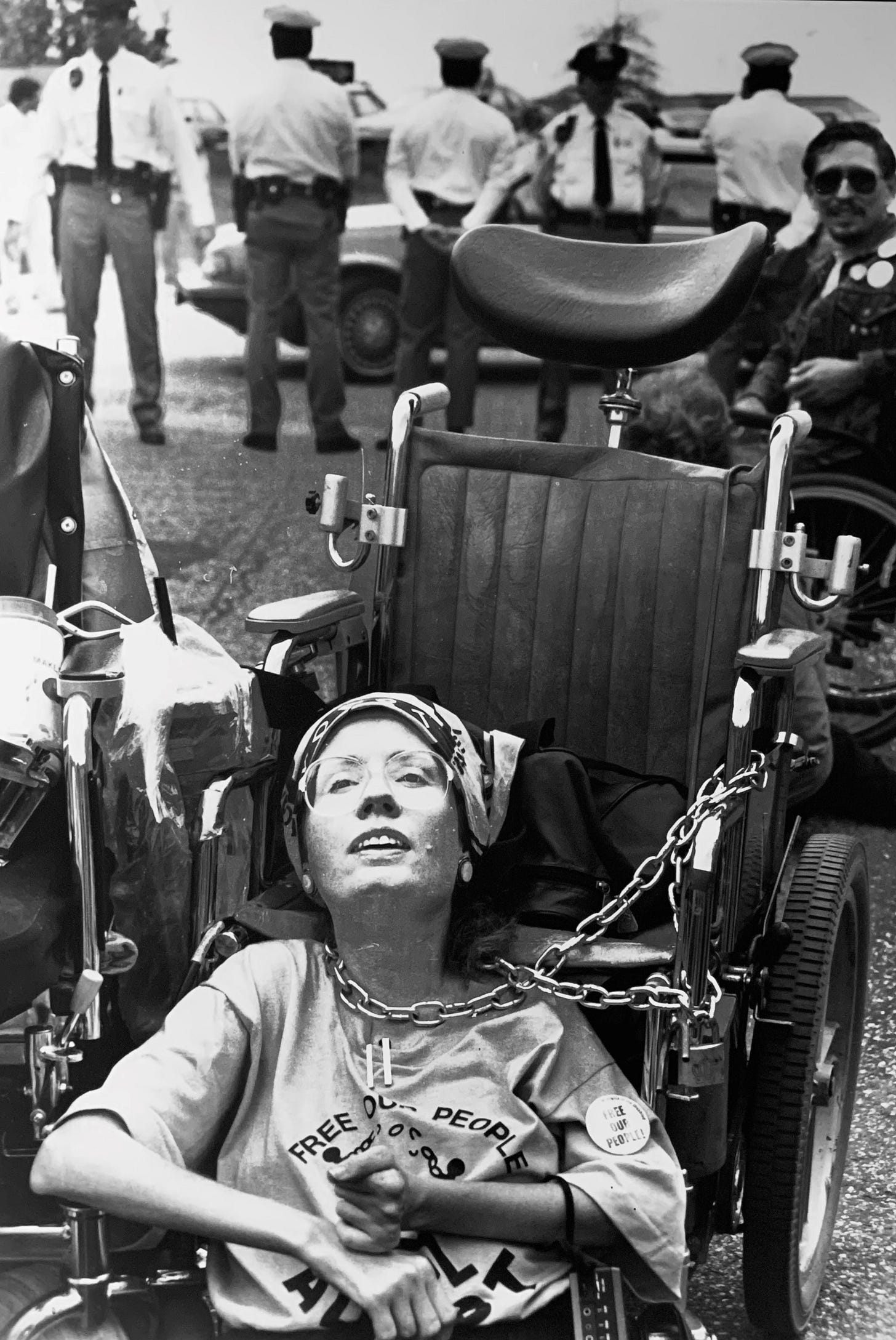 A white woman is on the ground in front of her manual wheelchair at a protest. She is attached to the chair by a metal chain wrapped around her neck. in the background, several police officers form a barrier.