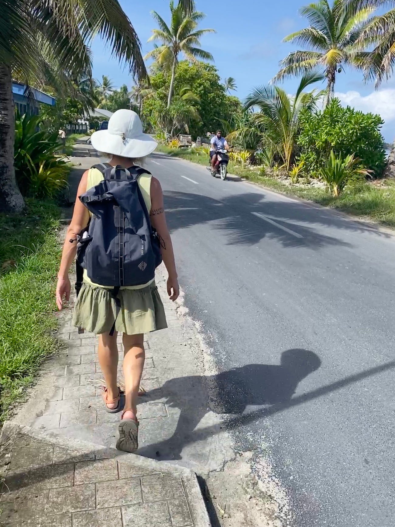 A woman wearing a skirt and a big sunhat walks along a paved road with tropical trees and plants alongside