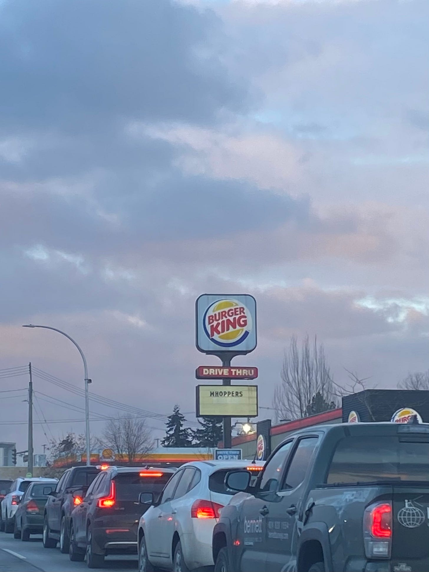 a line of cars at a red light. It’s nearing sunset and the sky is a bit cloudy, pale pinks and blue-grey. In the centre of frame is a large Burger King sign, the standard yellow circle with red text and a white background, the red drive thru sign above a yellowish marker with black letters that read ‘WHOPPERS’, slightly off centre.