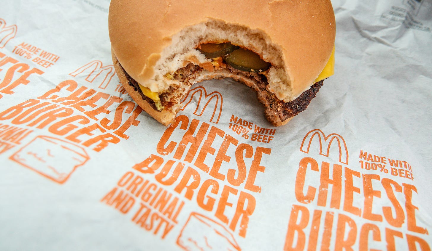 A close-up of a McDonald's cheeseburger, which has a bite taken out of it. The burger rests on the McDonalds wrapping, which says "made with 100% beef"
