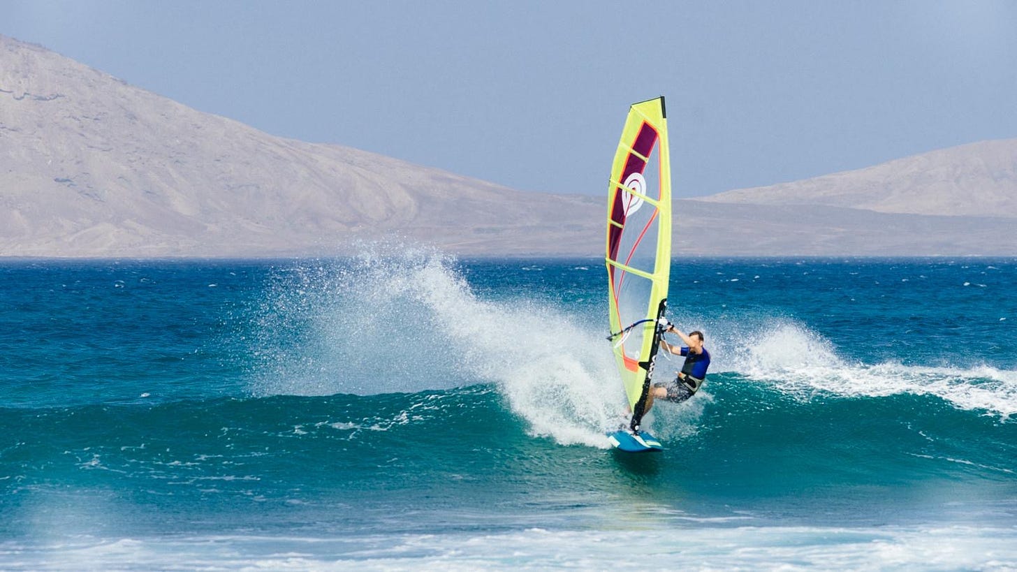 Mar com onda pequena e um rapaz numa prancha de windsurf. Atrás se vê uma região desértica.
