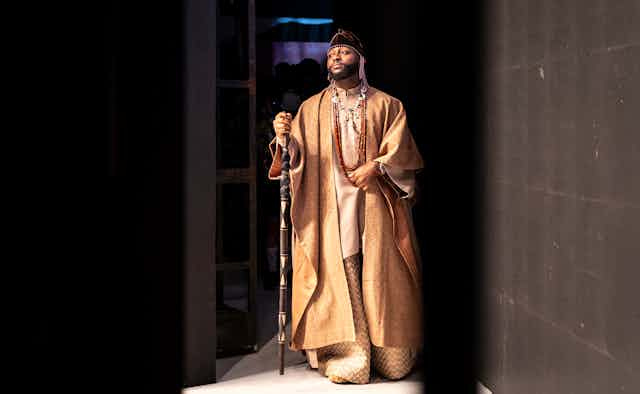 A man stands backstage behind curtains holding a walking stick and regally dressed in a flowing gold robe over loose-fitting gold top and trousers. A man stands backstage behind curtains holding a walking stick and regally dressed in a flowing gold robe over loose-fitting gold top and trousers.