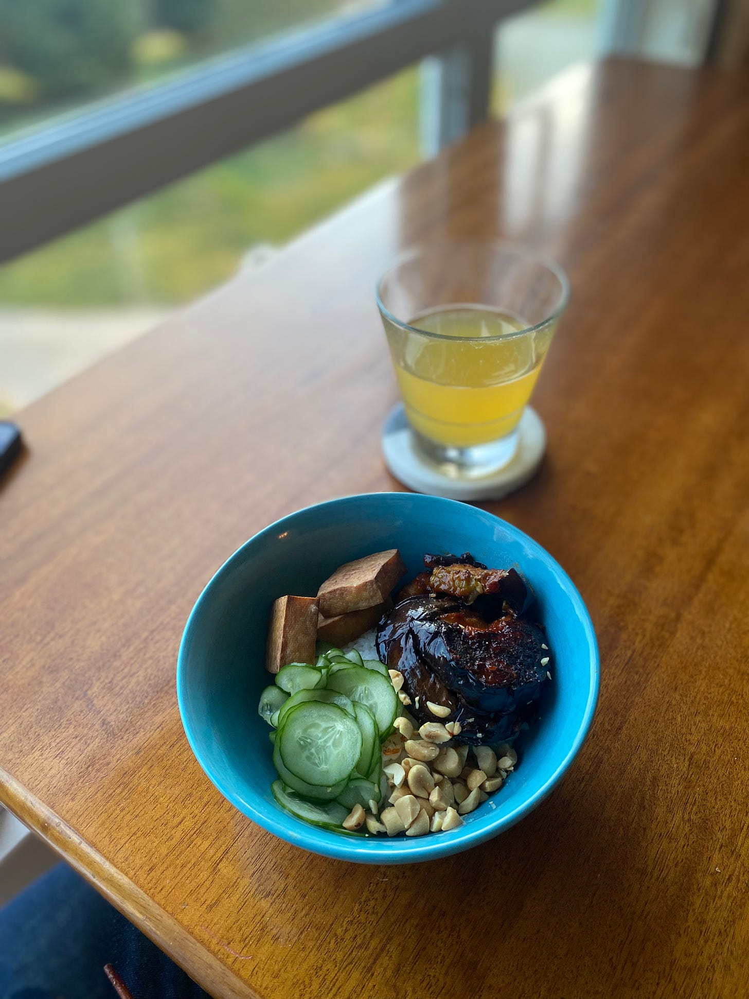 A blue bowl of rice topped with gochujang-glazed eggplant, peanuts, tofu, and quick-pickled cucumber. On a coaster is a glass of light-coloured beer.