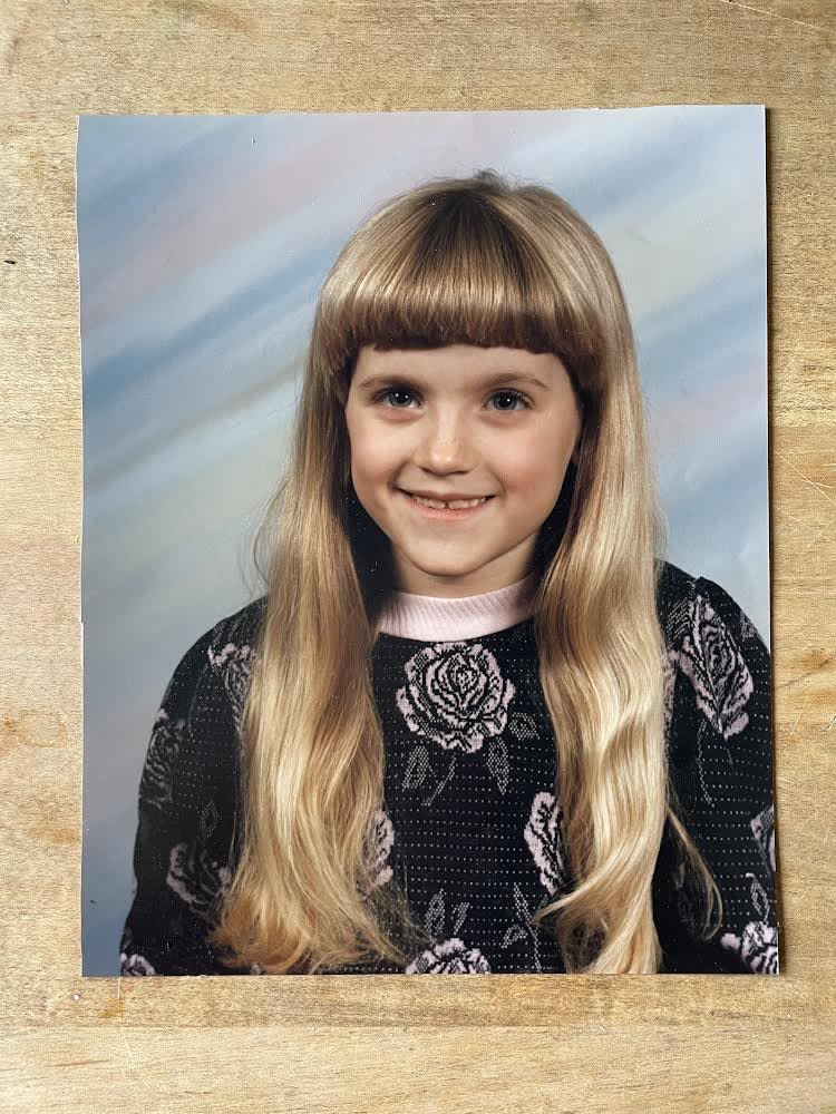 a small white child socialized as female with blonde bangs and long blonde hair is smiling at the camera