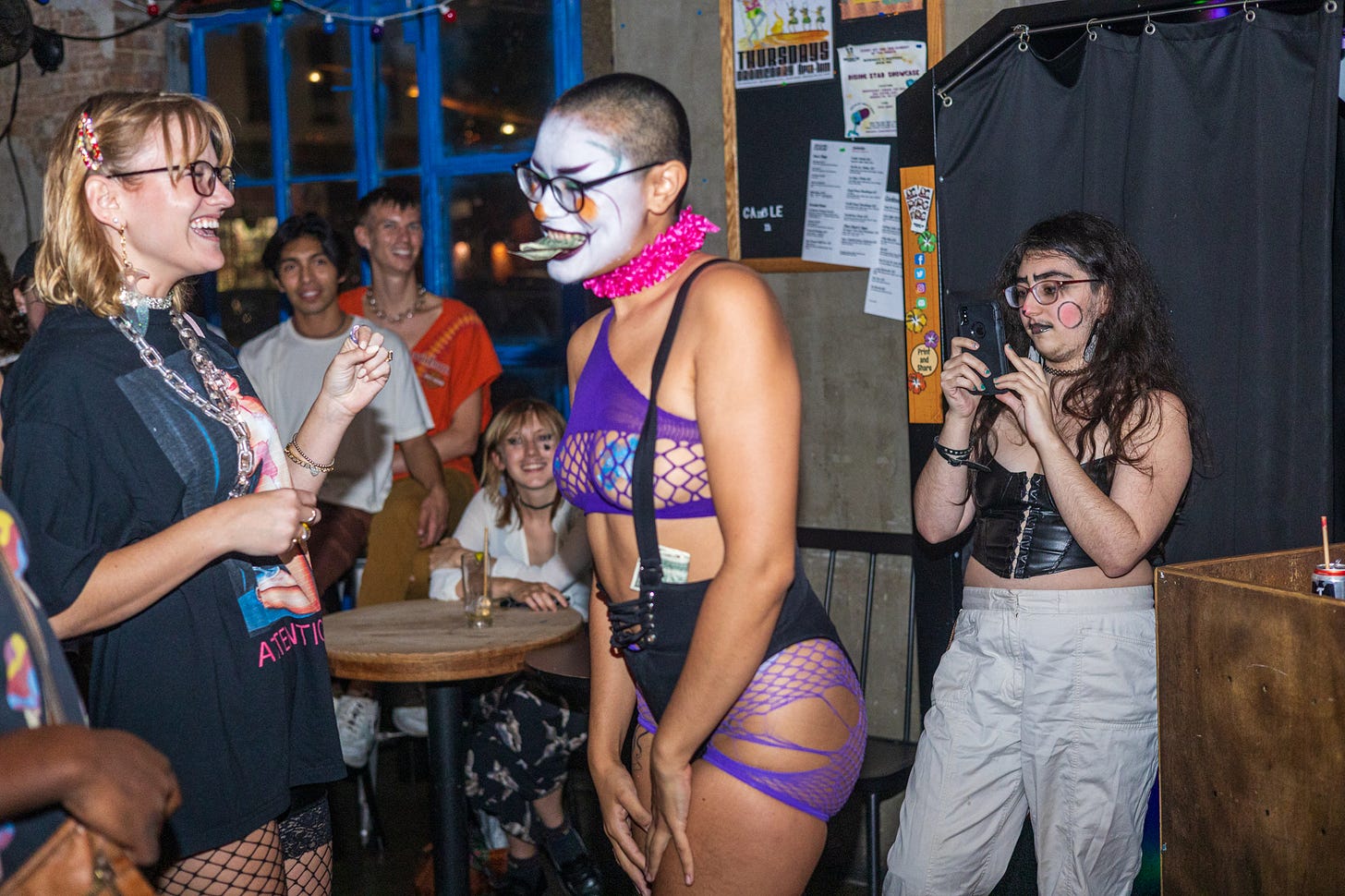 cochina divina, a nonbinary burlesque performer in white clown makeup and purple mesh lingerie holds dollar bill in mouth while audience surrounds them laughing and smiling
