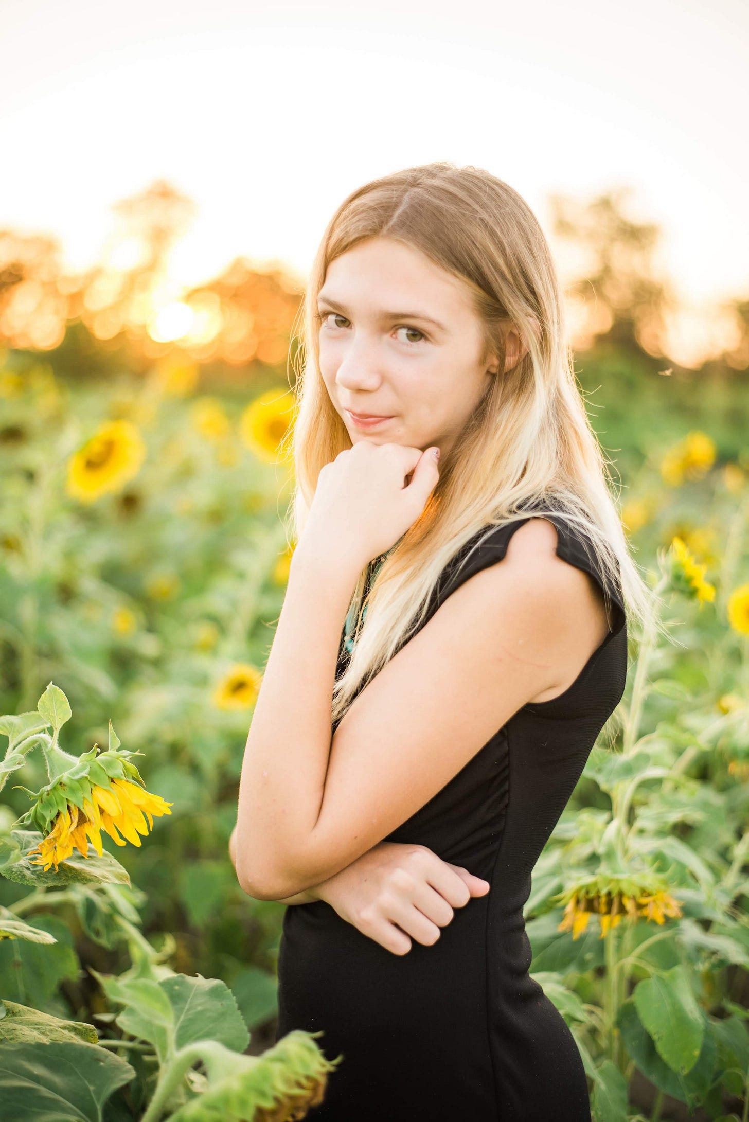 A blonde girl in a black dress, standing in a field of sunflowers, facing the viewer