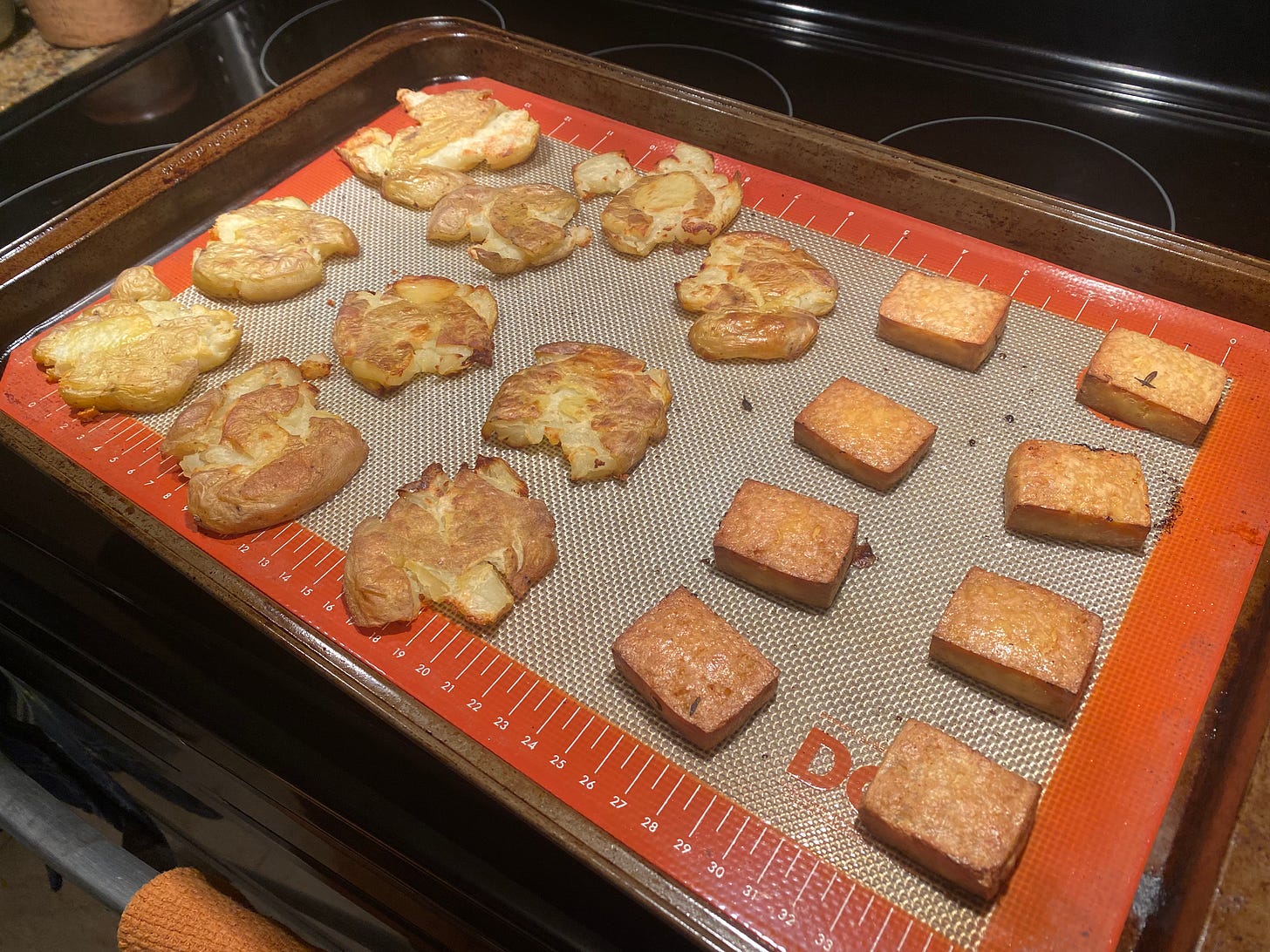 A baking sheet lined with a silpat. On the left are some smashed potatoes, skins split, edges and tops lightly browned. On the right are squares of marinated tofu, stained dark from the marinade and browned in the oven.
