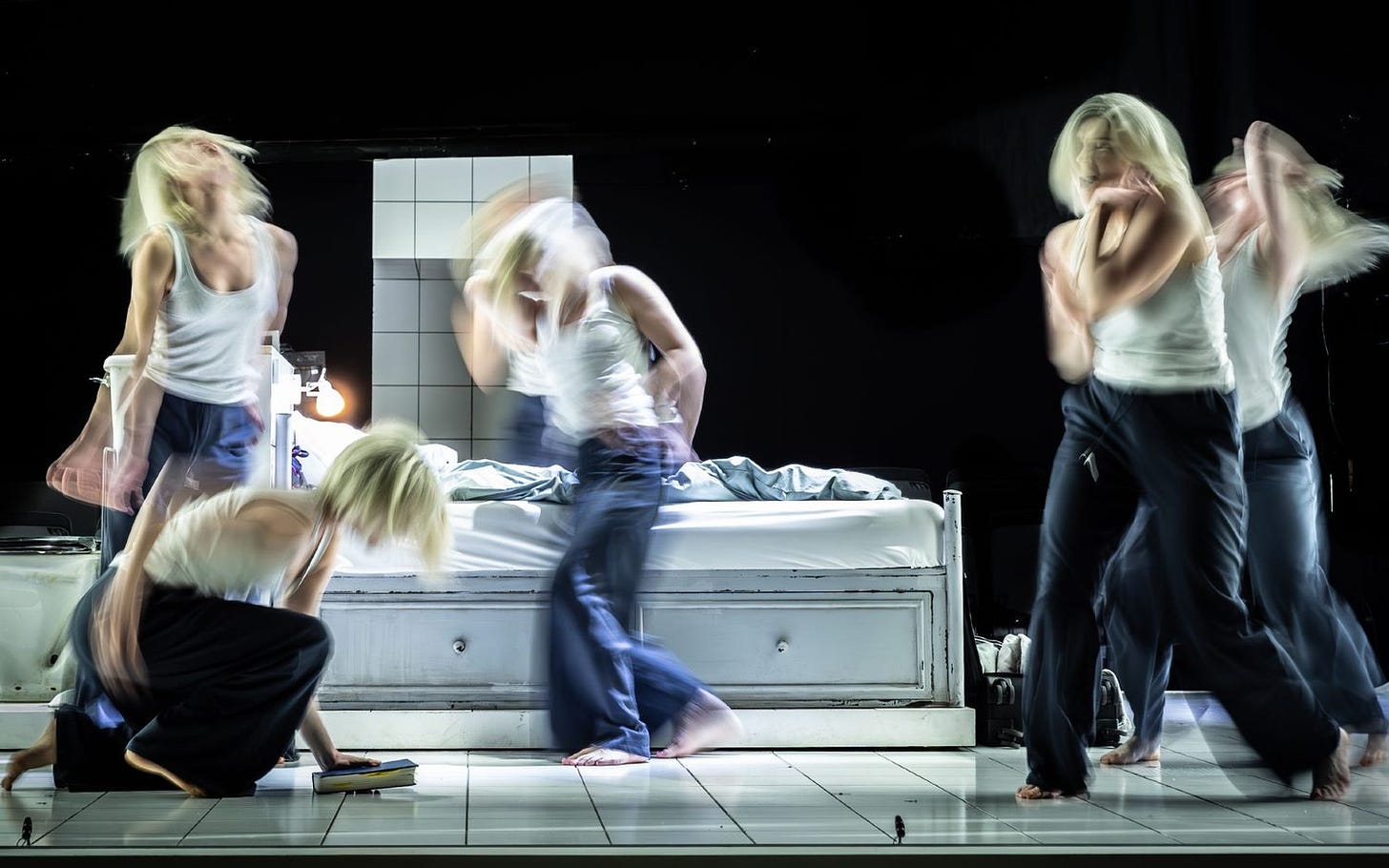 A dynamic theater scene featuring five actors in identical outfits and blond hair, blurred figures in expressive poses on a minimalist set, against a black background.