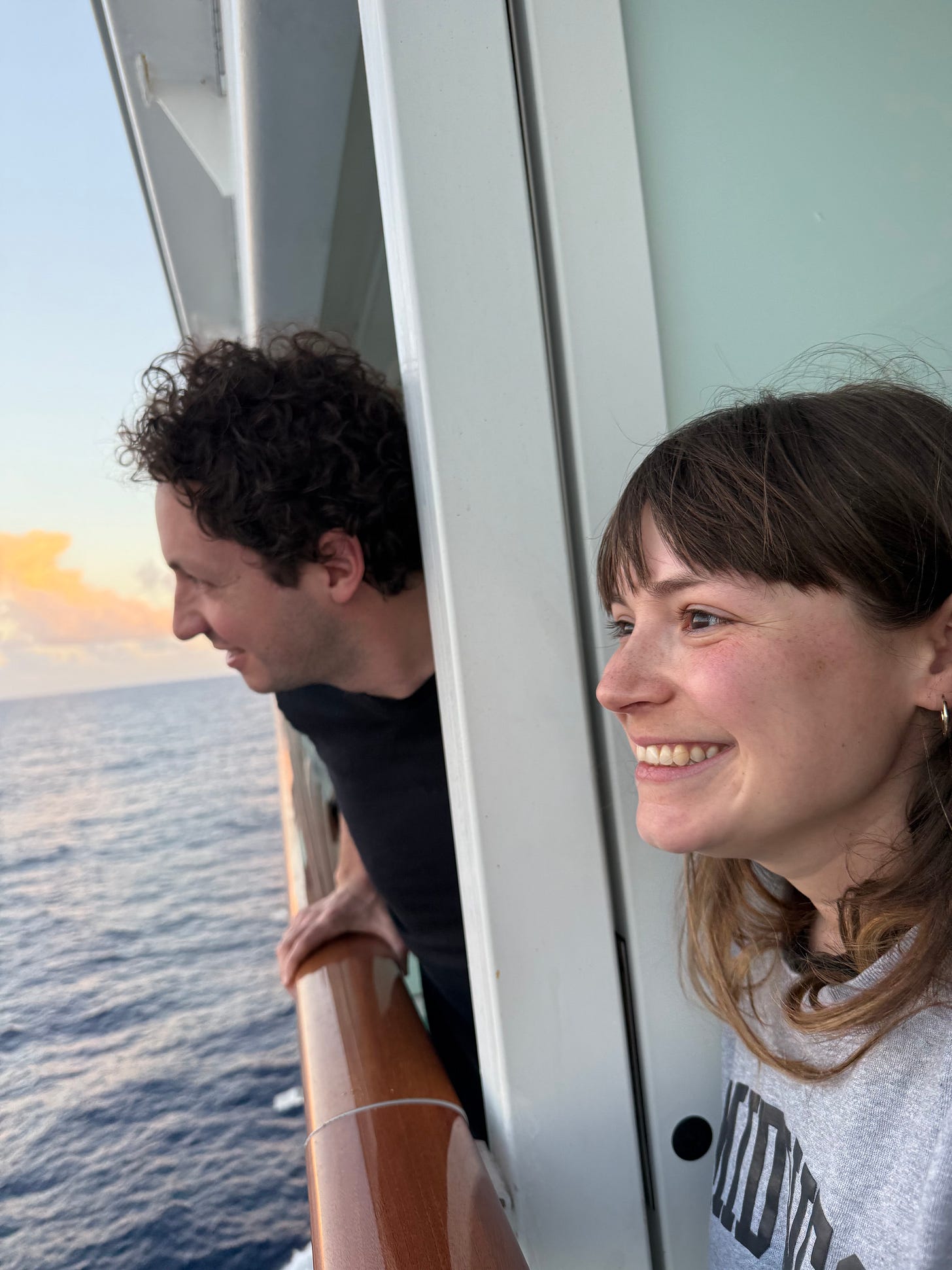 Two people smile and look at the ocean from a cruise ship stateroom balcony.