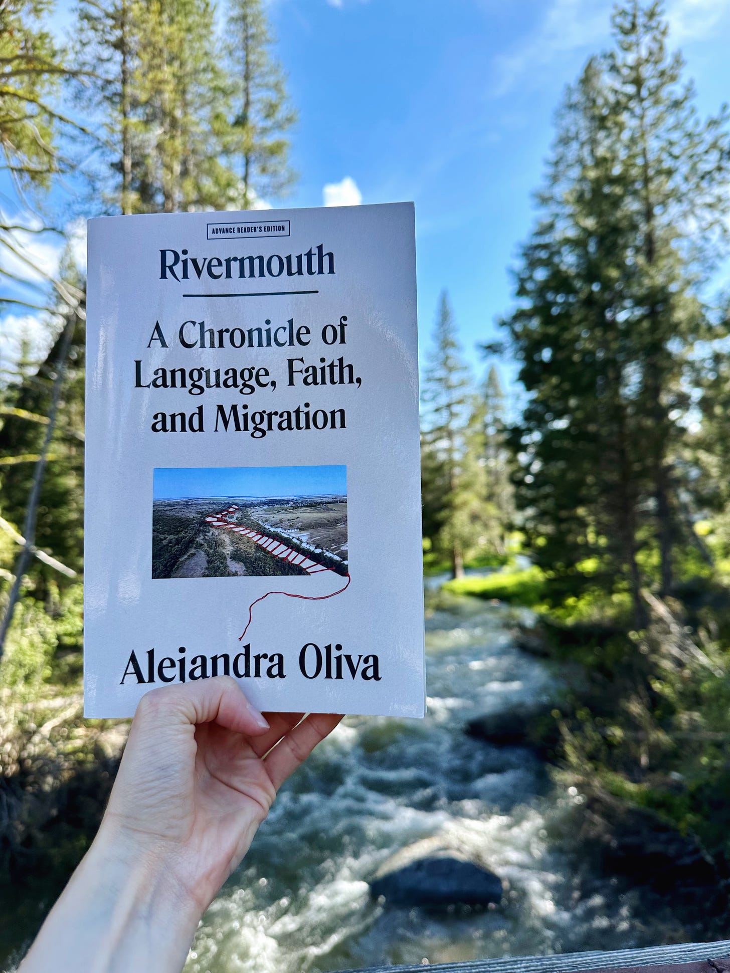 A white hand holds up a copy of Rivermouth in front of a river, green trees, and a blue sky
