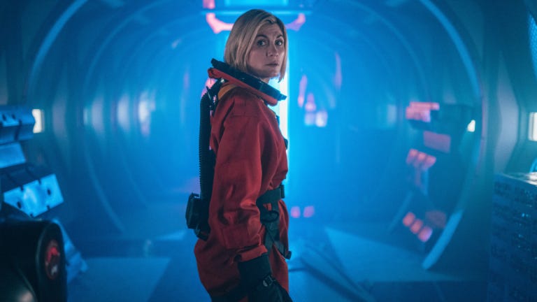 This image shows the Thirteenth Doctor (played by Jodie Whittaker) standing in a space bullet train's cargo carriage wearing an orange space suit and looking back at camera. Blue light suffuses the frame. This image shows the Thirteenth Doctor (played by Jodie Whittaker) standing in a space bullet train's cargo carriage wearing an orange space suit and looking back at camera. Blue light suffuses the frame.