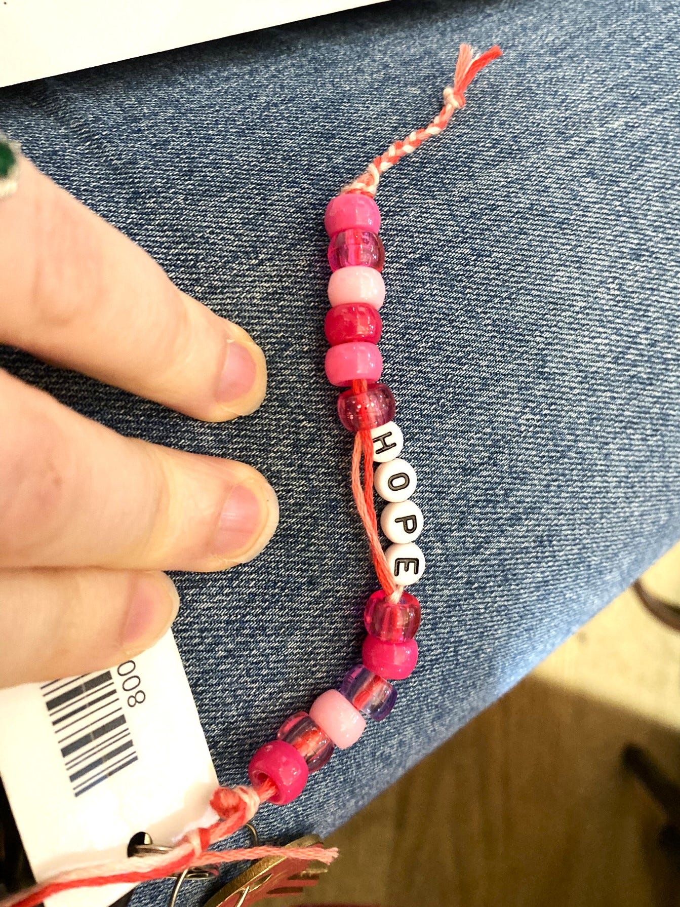 A pink friendship bracelet is laid out on a leg (wearing jeans). In the middle of the bracelet are four beads that spell out HOPE.