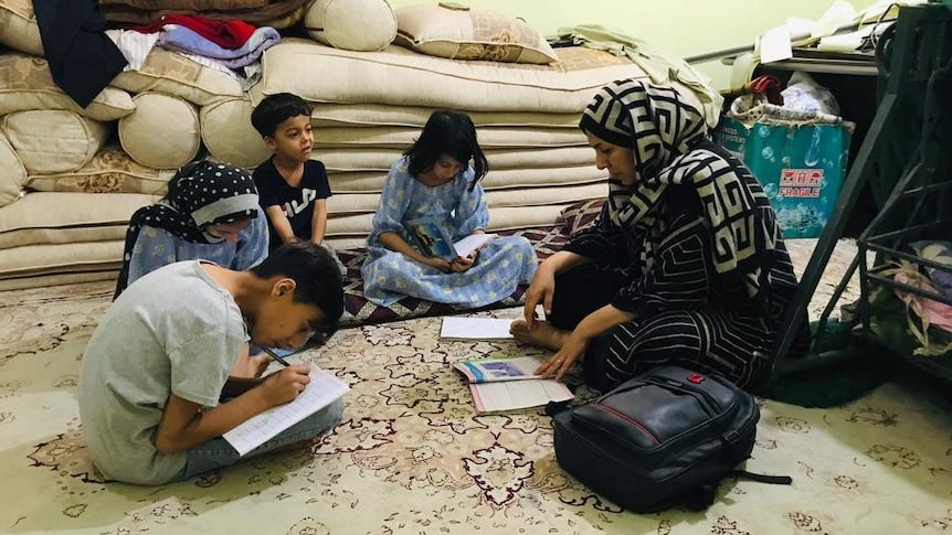 A mother sits on a rug with her four of her children as they read and write on white sheets of paper.