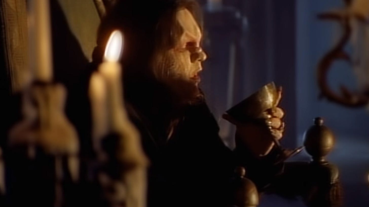 Still from Meatloaf's music video for I Would Do Anything For Love (But I Won't Do That) where Meatloaf in beast/vampire creature makeup stares into a goblet while sitting in a room lit by candles.