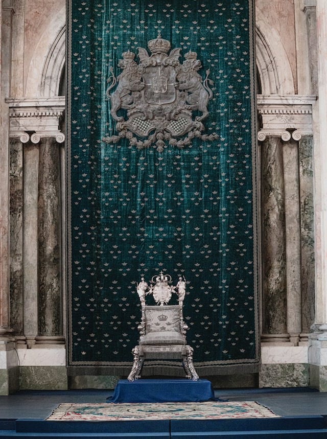 An ornate but small silver throne with gray cushions in front of a long deep green tapestry. High above the throne is an ornate gray insignia. The throne sits atop a rectangular shaped block, covered in deep blue fabric, which itself set behind a rug.