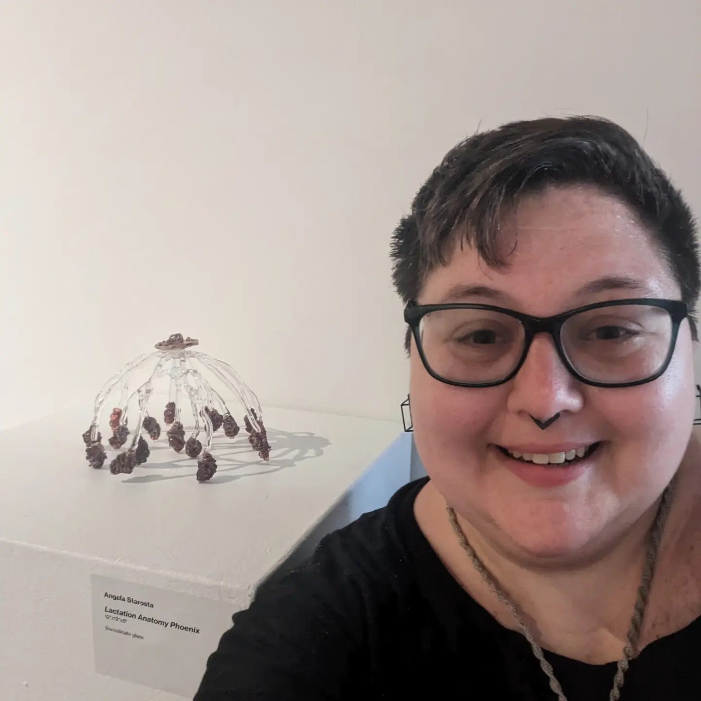 Angela, pictured with short dark hair, dark acrylic glasses, and septum ring, poses for a selfie with a glass sculpture in a gallery. The sculpture is the lactation anatomy of the breast (dome shaped with spider-leggy ducts running from mammary) and sits on a pedestal with her name and information on it.