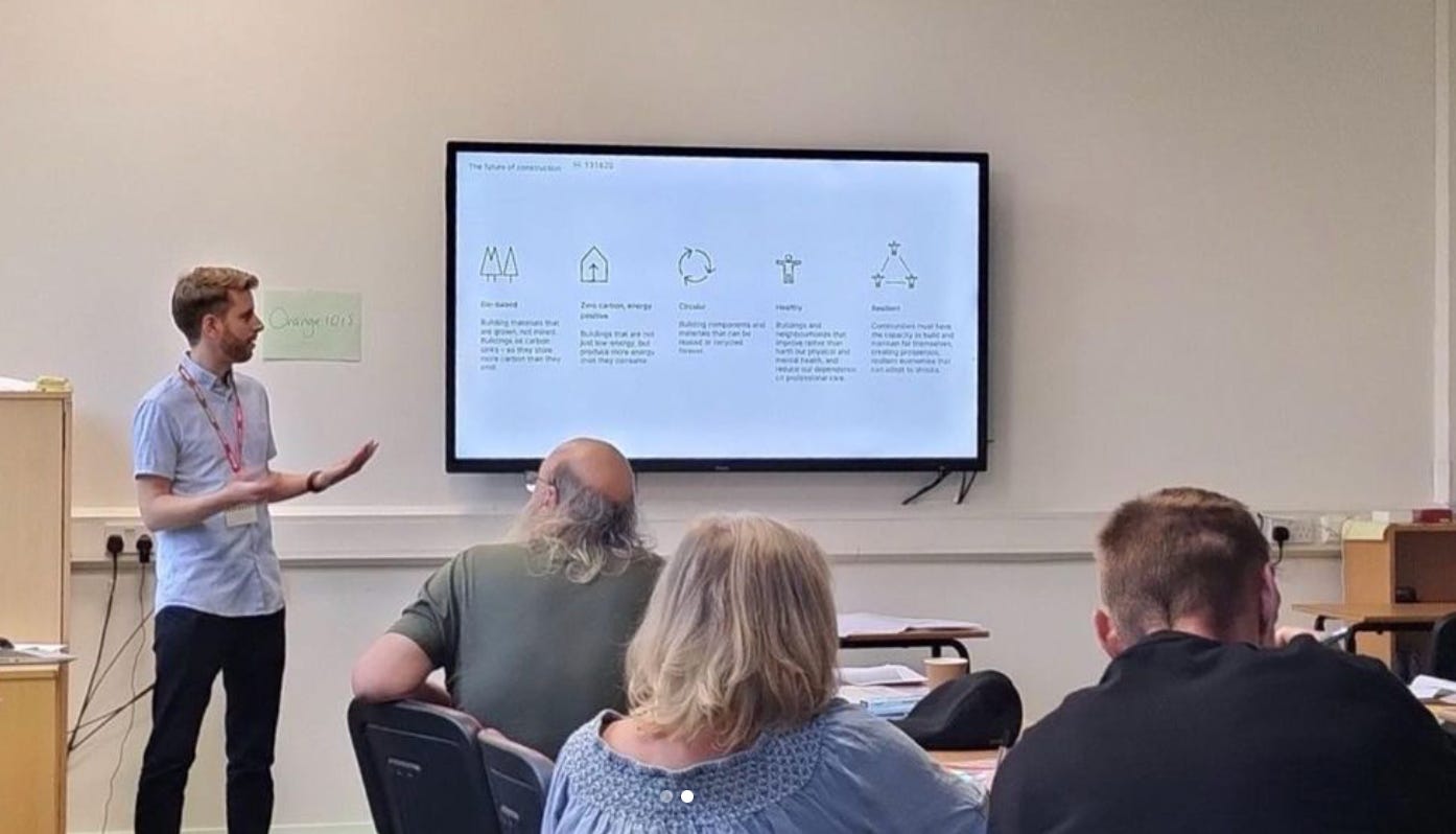 A man stands in front of a room, delivering a talk to people and showing a presentation on a large tv screen.