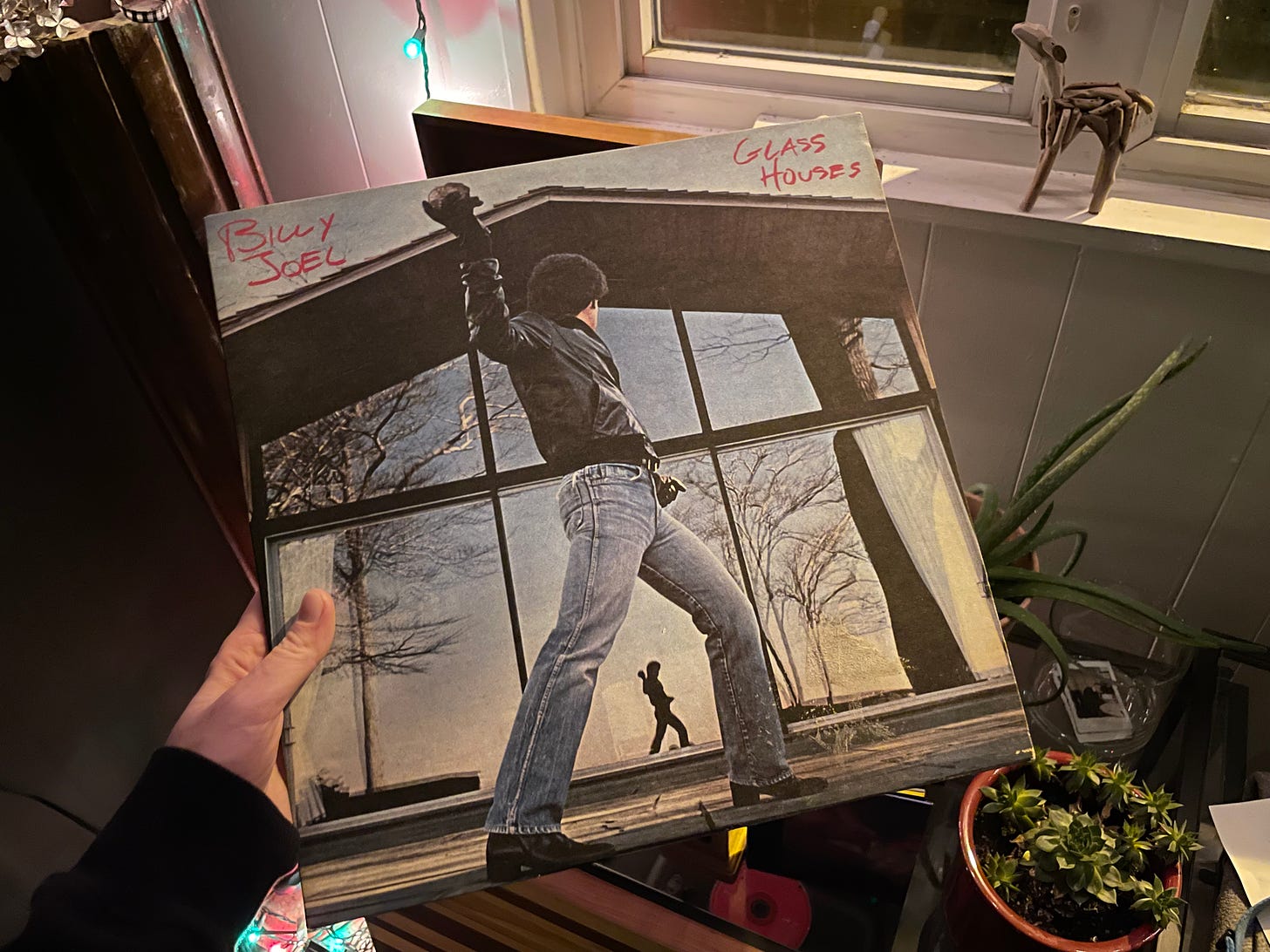Photograph of a record of Billy Joel's Glass Houses, which features a man in a leather jacket in jeans throwing a rock at a large window.