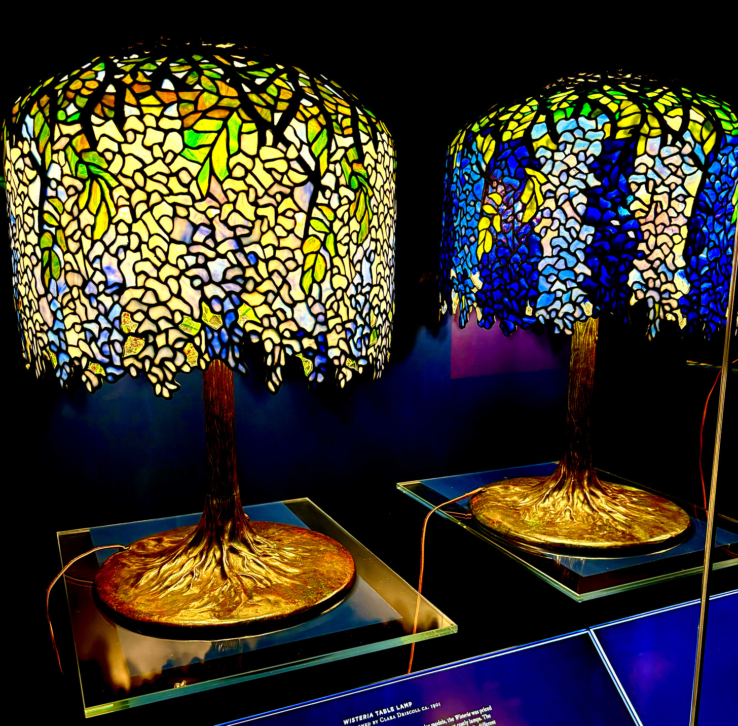 Two wisteria lamp shades and engraved bases. One is striped darker and lighter blue. The one on the left is more blue interspersed with green glass.