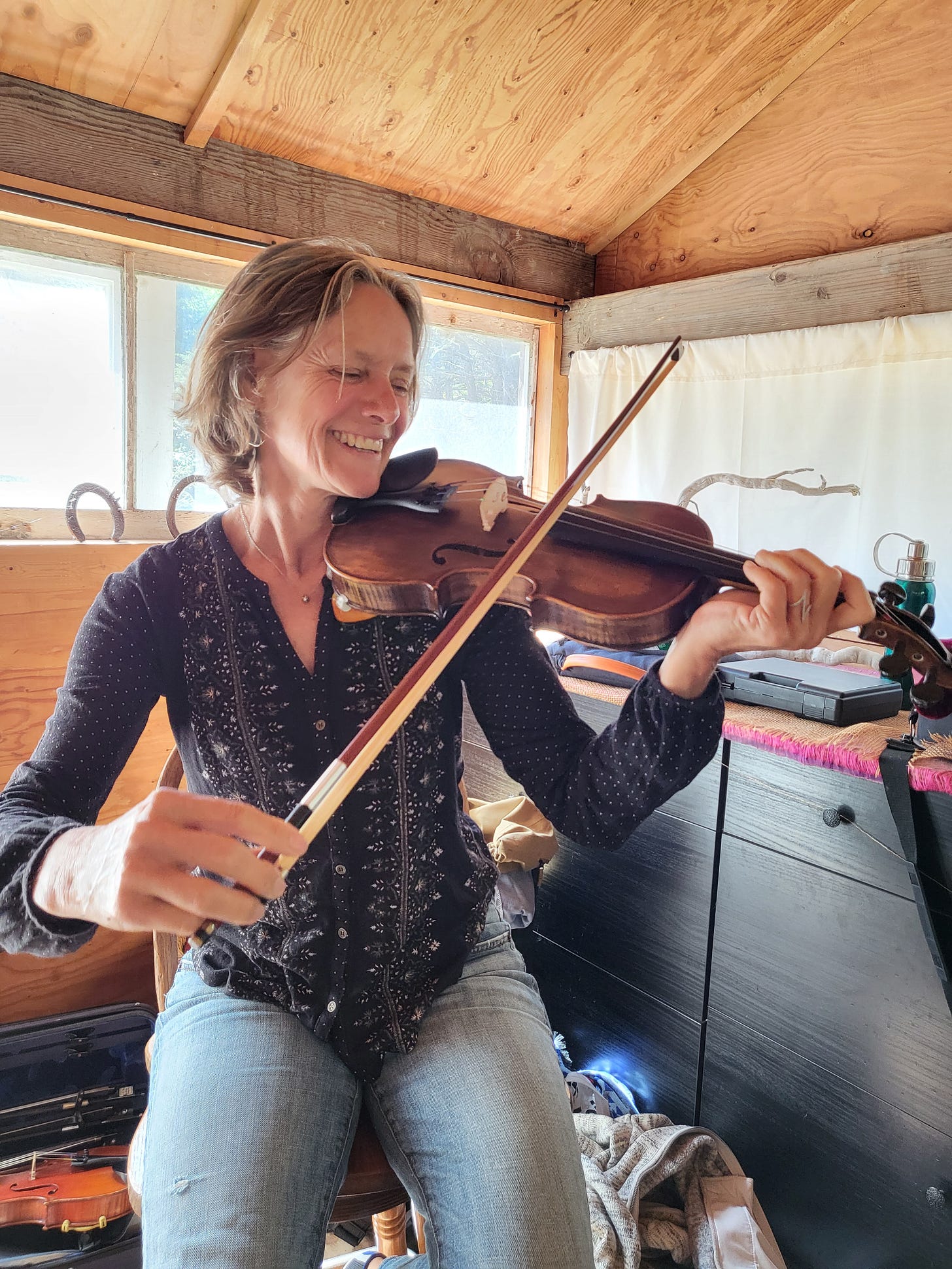 Melanie sitting on a stool in a rustic shed, playing fiddle and smiling