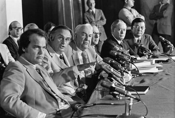 Several men in suits and ties, including Mr. Weicker, seated at a table with many microphones on it. They all have very serious expressions on their faces.