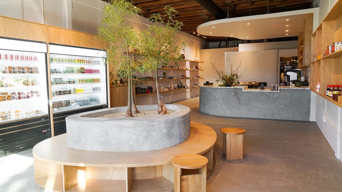 Inside Suá Superette in Larchmont Village with a concrete center area with live trees, wood bench seating, fridges, and a counter to the back.